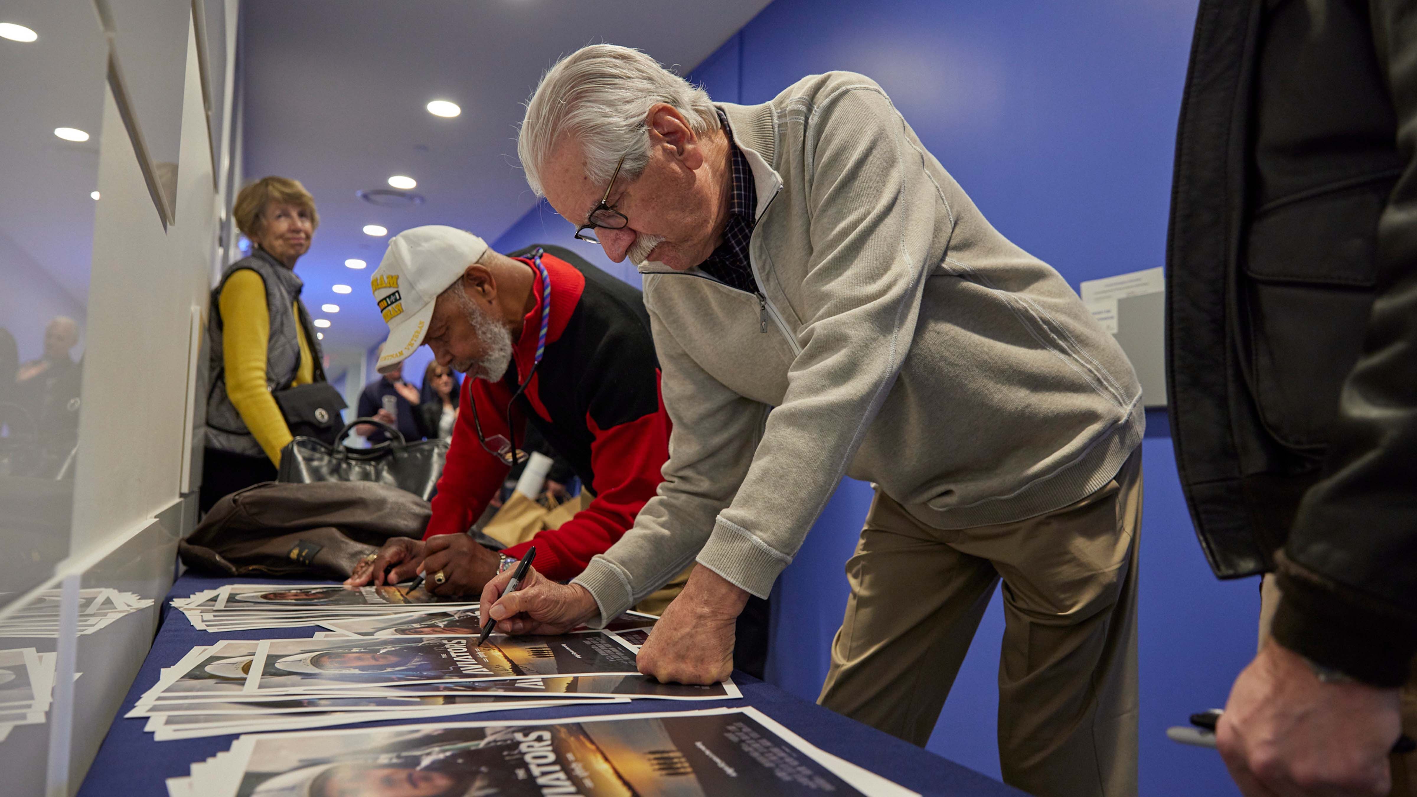 Two male army veterans, Elester 'Hollywood' Latham and Bill 'Offender' Moir, signing posters for the premier of the film they are featured in.